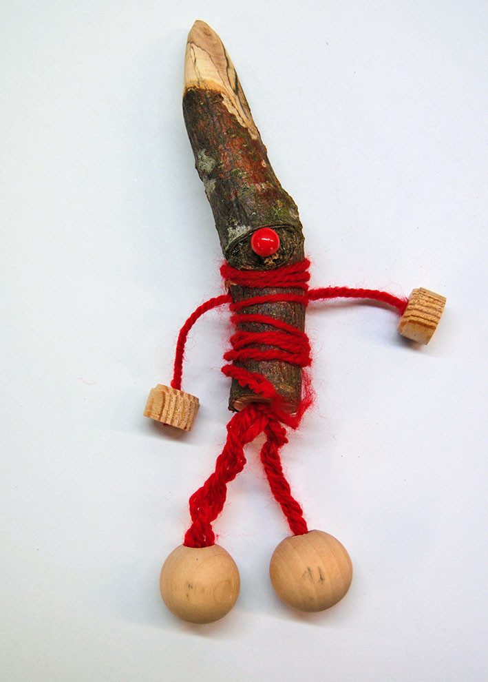 Holzfigur mit Stock als Körper, kleiner roter Perle als Nase und Holzkugeln an Wolle als Arme und Beine