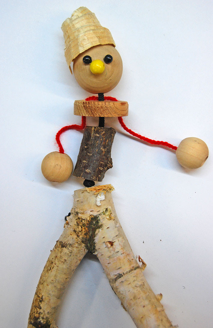 Holzfigur mit Stöcken als Beine und Körper, Holzperlen an Wolle als Arme und großer Holzkugel als Kopf mit Augen und Nase aus Perlen