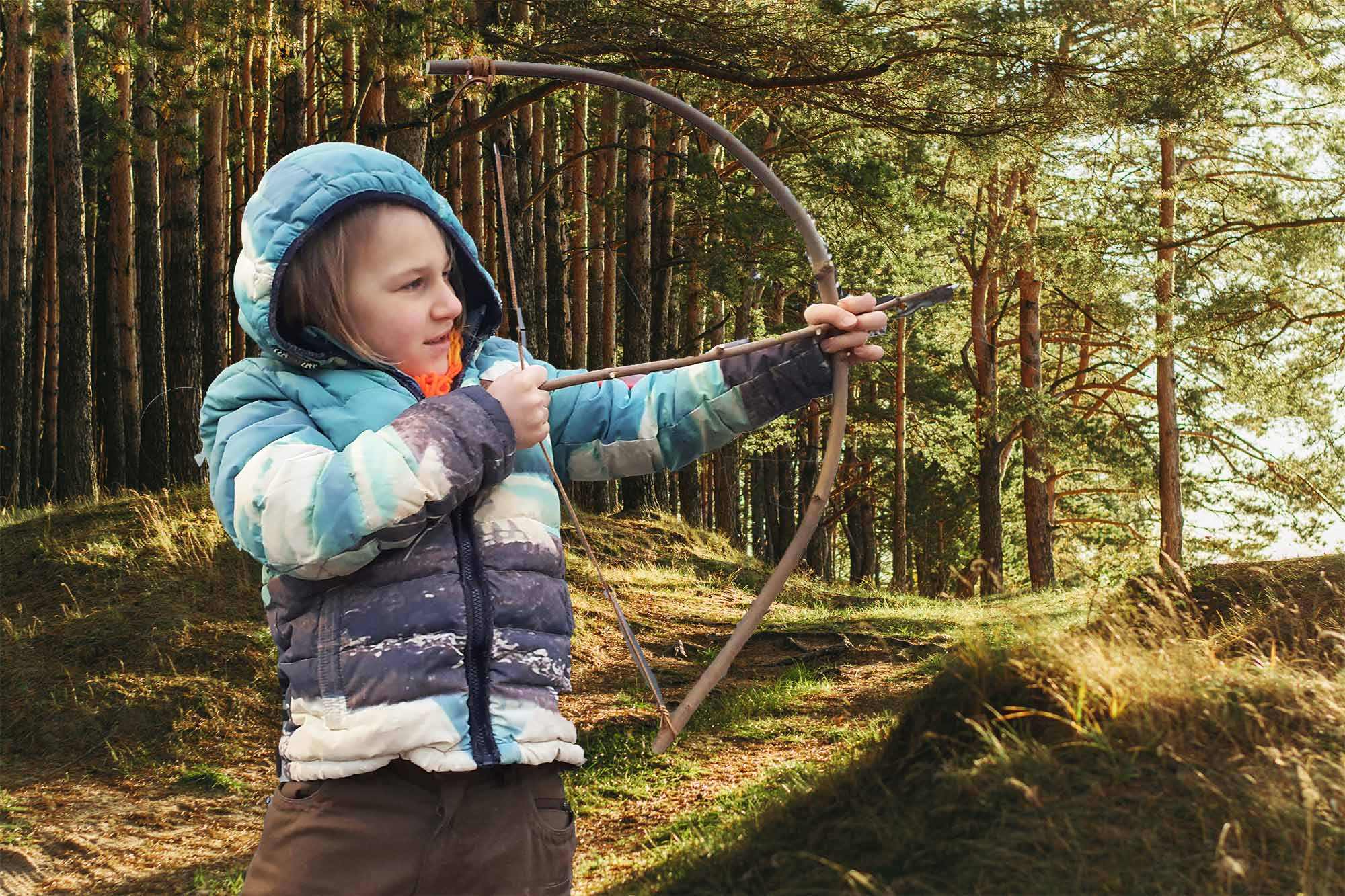 Kind mit selbstgebauten Pfeil und Bogen beim Bogen spannen vor Waldhintergrund
