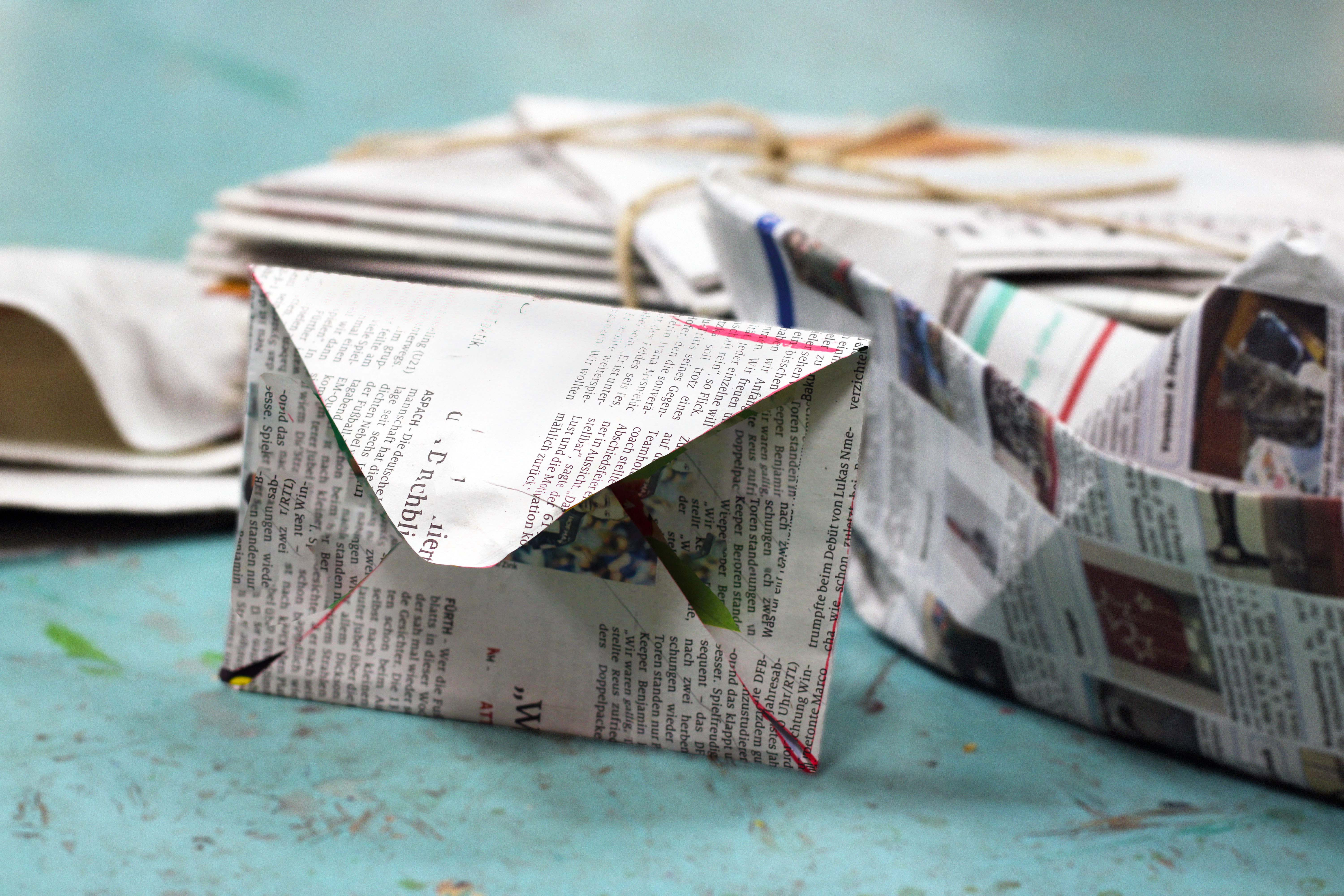 Briefumschlag aus Zeitungspapier, dahinter ein gefaltetes Schiffchen aus Zeitungspapier vor einem Hintergrund aus einem Zeitungsstapel auf blauem Boden