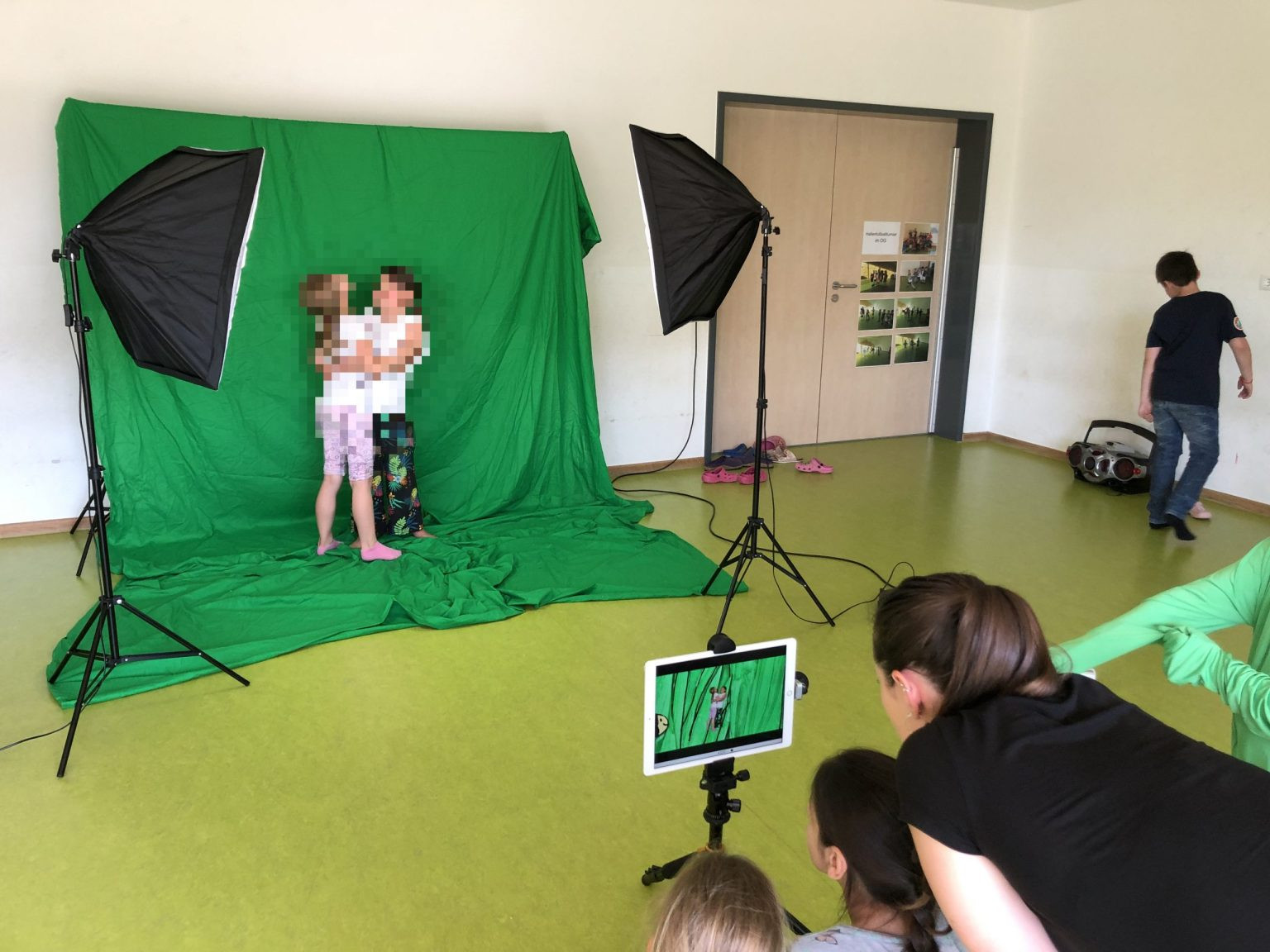 Zwei sich umarmende Kinder vor einer grünen Stoffwand, beleuchtet mit Softboxen, im Vordergrund weitere Kinder und Dozentin, die über ein Tablet die Szenerie begutachten