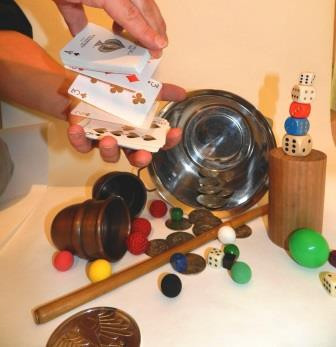 verschiedene Zaubermaterialien wie Würfel, Becher, verschieden farbige Kugeln, Münzen - oben rechts Kinderhände, die Spielkarten mischen