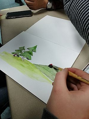 Kind malt mit Aquarell-Farben eine Wiese mit zwei Tannenbäumen