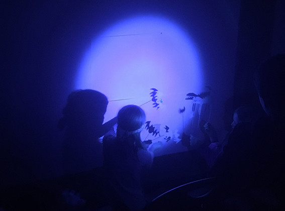 nachtblauer Raum mit Lichtkegel vor dem man Kind sieht, dass mit Schattenwurf von Papierfiguren experimentiert
